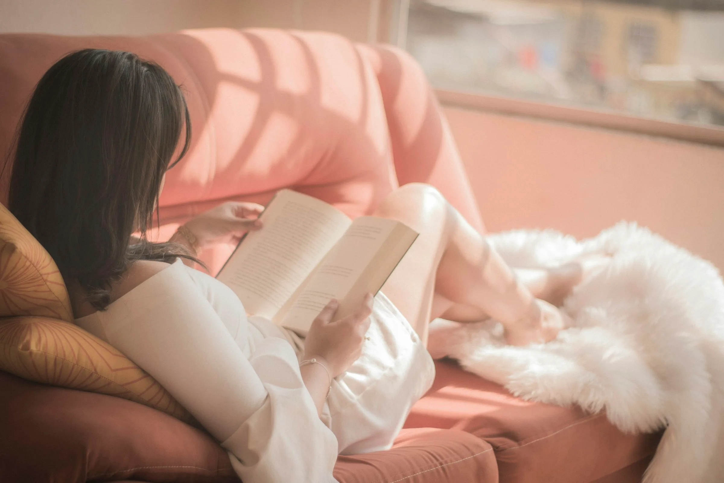 Jeune femme qui lit un livre, allongée au soleil sur un canapé rose